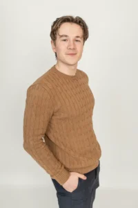 Justdata ICT, Een jonge man met een lichte huid en kort bruin haar staat tegen een effen lichte achtergrond, draagt een bruine kabeltrui en een donkere broek, met een hand in zijn zak en een lichte glimlach op zijn gezicht.